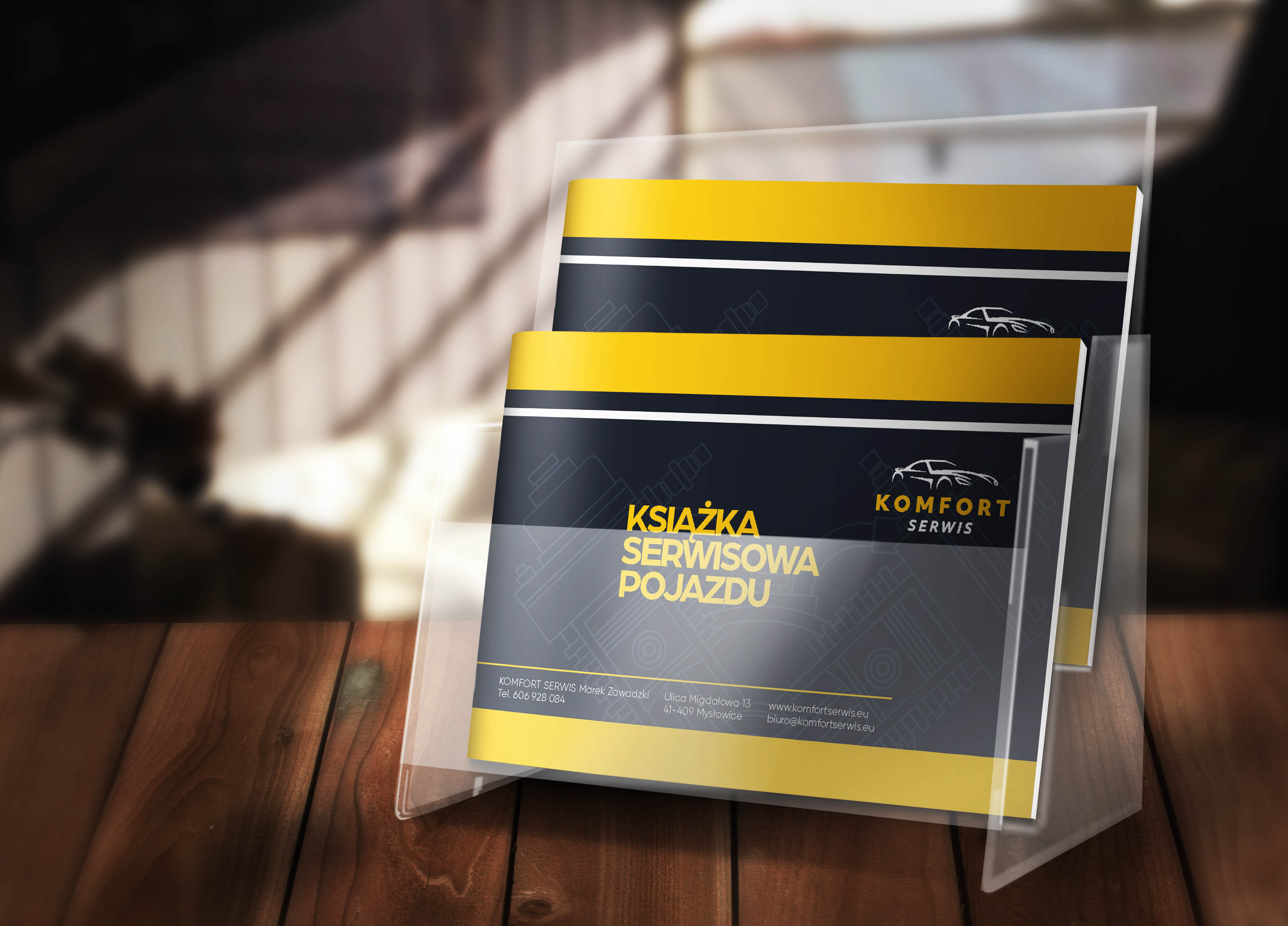 Dwa foldery reklamowe serwisu samochodowego 'Komfort Serwis' w przezroczystym stojaku na drewnianym blacie. Foldery w kolorystyce szaro-żółtej, z napisem 'Książka Serwisowa Pojazdu' i schematycznym...