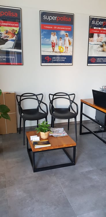 Poczekalnia z dwoma czarnymi krzesłami o ażurowym oparciu, stolikiem kawowym z gazetami i rośliną doniczkową, oraz plakatami firmy ubezpieczeniowej Superpolisa na ścianie.