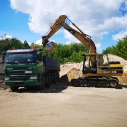 Żółta koparka CAT 318B L nabiera łyżką piasek i wsypuje go na pakę zielonej ciężarówki DAF CF, w tle drzewa i błękitne niebo z chmurami.