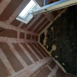 Poddasze w trakcie prac wykończeniowych: widoczne płyty kartonowo-gipsowe na ścianach, częściowo pokryte warstwą szpachli, okno dachowe i fragment muru z cegły. Na górze fragment drabiny.