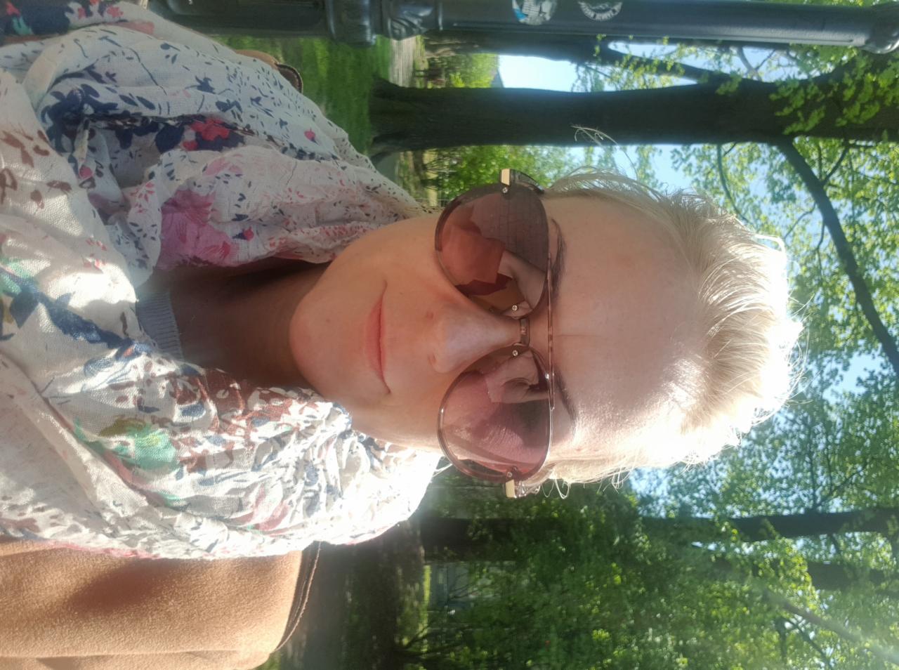 Portret kobiety w okularach przeciwsłonecznych, na tle zieleni parku i fragmentu metalowej konstrukcji ławki.