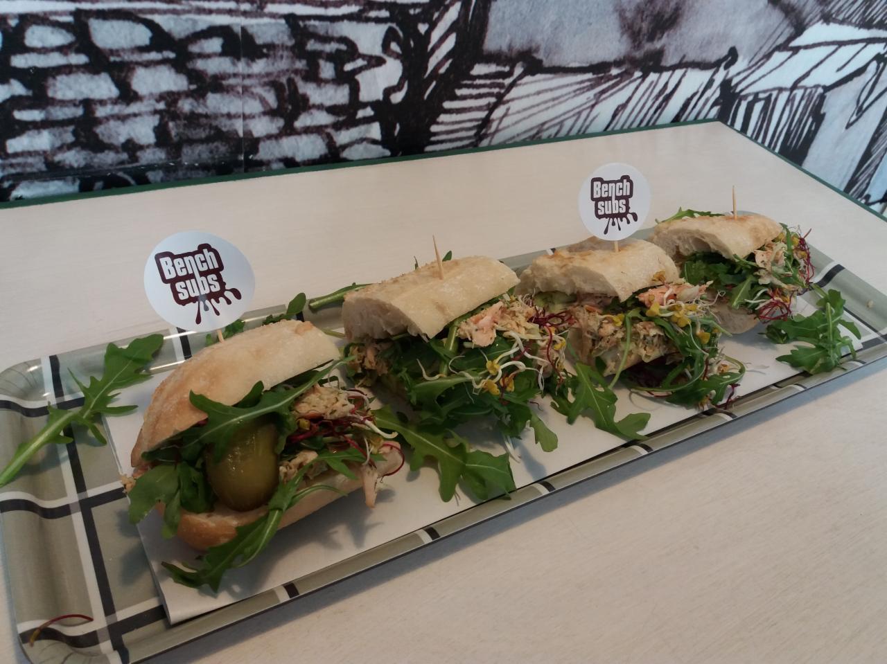 Cztery kanapki 'Bench Subs' na tacy z rukolą i kiełkami, każda z innym dodatkiem, na tle grafiki przypominającej mur z cegieł.
