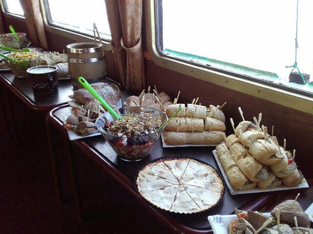 Stół z różnorodnymi przekąskami: mini kanapki z wykałaczkami, sałatki w miskach z zielonymi łyżkami, tarta na słodko, ustawione przy oknie w pomieszczeniu o ciemnych ścianach.