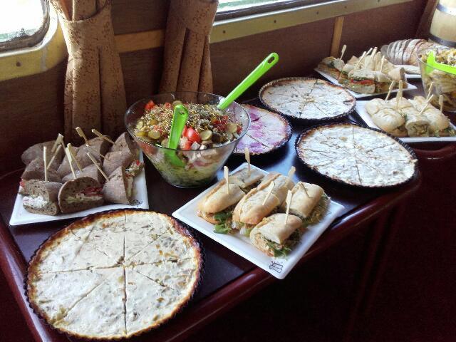 Stół z różnorodnym cateringiem: mini kanapki na szpilkach, sałatka w szklanej misie z zielonymi sztućcami, tarty podzielone na porcje, oraz roladki na białych talerzach. Widoczne zasłony w tle...