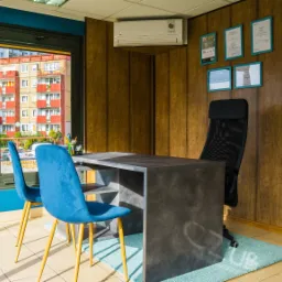 Biuro agencji nieruchomości z widokiem na osiedle mieszkaniowe, biurko z szarą fakturą, dwa błękitne krzesła, certyfikaty na ścianie i klimatyzator.