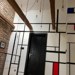 Wnętrze w stylu Mondriana z drewnianymi belkami stropowymi, czarnymi liniami na białych ścianach z akcentami czerwieni i błękitu, oraz wiszącymi żar&oacute;wkami na kablach.