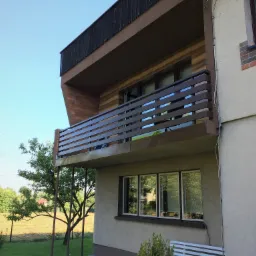 Balkon z brązową, poziomą balustradą na tle błękitnego nieba i zieleni ogrodu, widok z dołu.