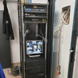Szafa rack z zainstalowanym sprzętem sieciowym: switche, patch panele, rejestrator monitoringu wizyjnego z podglądem z kamer na monitorze. Widoczne okablowanie i przewody. Ściana z otworem na kable.