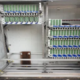 Wewnętrzny widok szafy telekomunikacyjnej z panelami krosowniczymi, widoczne liczne złącza światłowodowe z niebieskimi zabezpieczeniami, kable prowadzone wzdłuż metalowej ramy konstrukcyjnej.