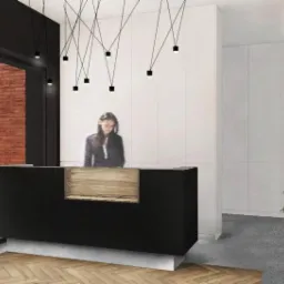 Nowoczesna recepcja w biurze z czarnym blatem i drewnianym elementem, oświetlona wiszącymi lampami, z widoczną pracownicą i rośliną doniczkową w tle.