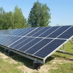 Rząd paneli słonecznych na metalowej konstrukcji, ustawiony na trawiastym terenie z drzewami w tle pod błękitnym niebem.