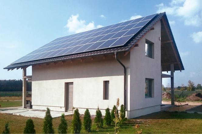 Dom jednorodzinny z panelami słonecznymi na dachu, widok z boku na tle błękitnego nieba z chmurami, małe tuje w ogródku.