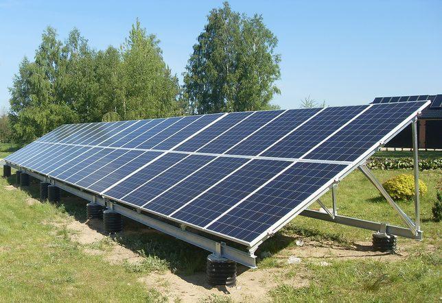 Rząd paneli słonecznych na metalowej konstrukcji, ustawiony na trawiastym terenie z drzewami w tle pod błękitnym niebem.