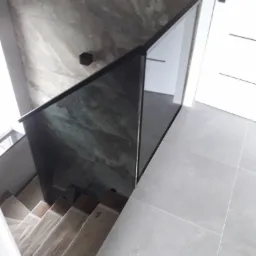 Szklana balustrada z czarnymi profilami zabezpieczająca schody drewniane, widok z góry, ściana z efektem marmuru i białe drzwi w tle.