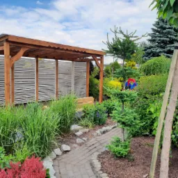 Zadaszona konstrukcja ogrodowa z drewnianymi słupami i białymi panelami żaluzjowymi, otoczona bujną roślinnością i ścieżką z kostki brukowej.