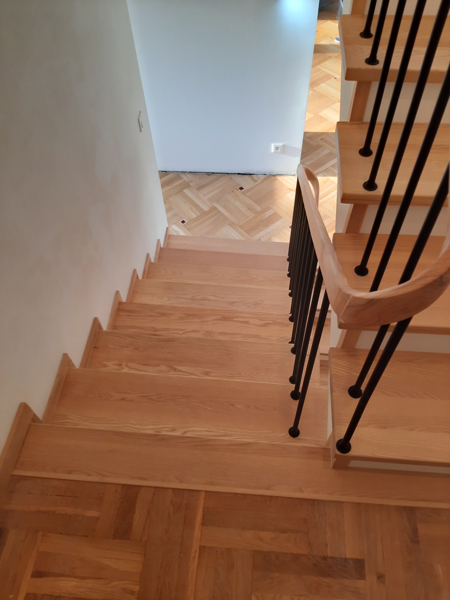 Schody drewniane z jasnego drewna, z czarnymi, metalowymi poręczami, widok z góry na dół, parkiet w jodełkę na podeście.