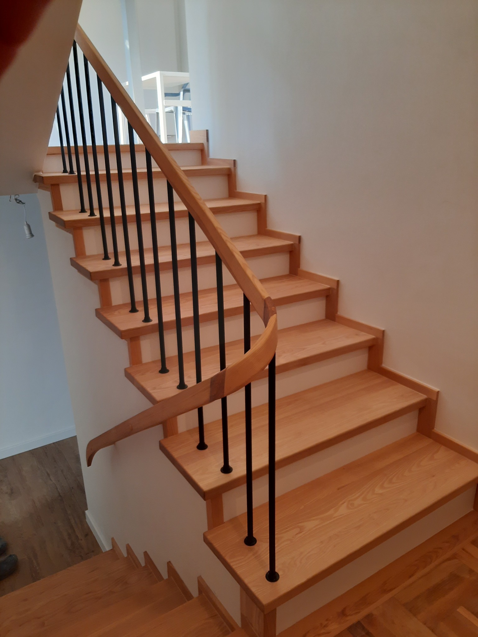 Jasne, drewniane schody z czarnymi, metalowymi poręczami w nowoczesnym wnętrzu. Widoczne delikatne usłojenie drewna i minimalistyczny design.