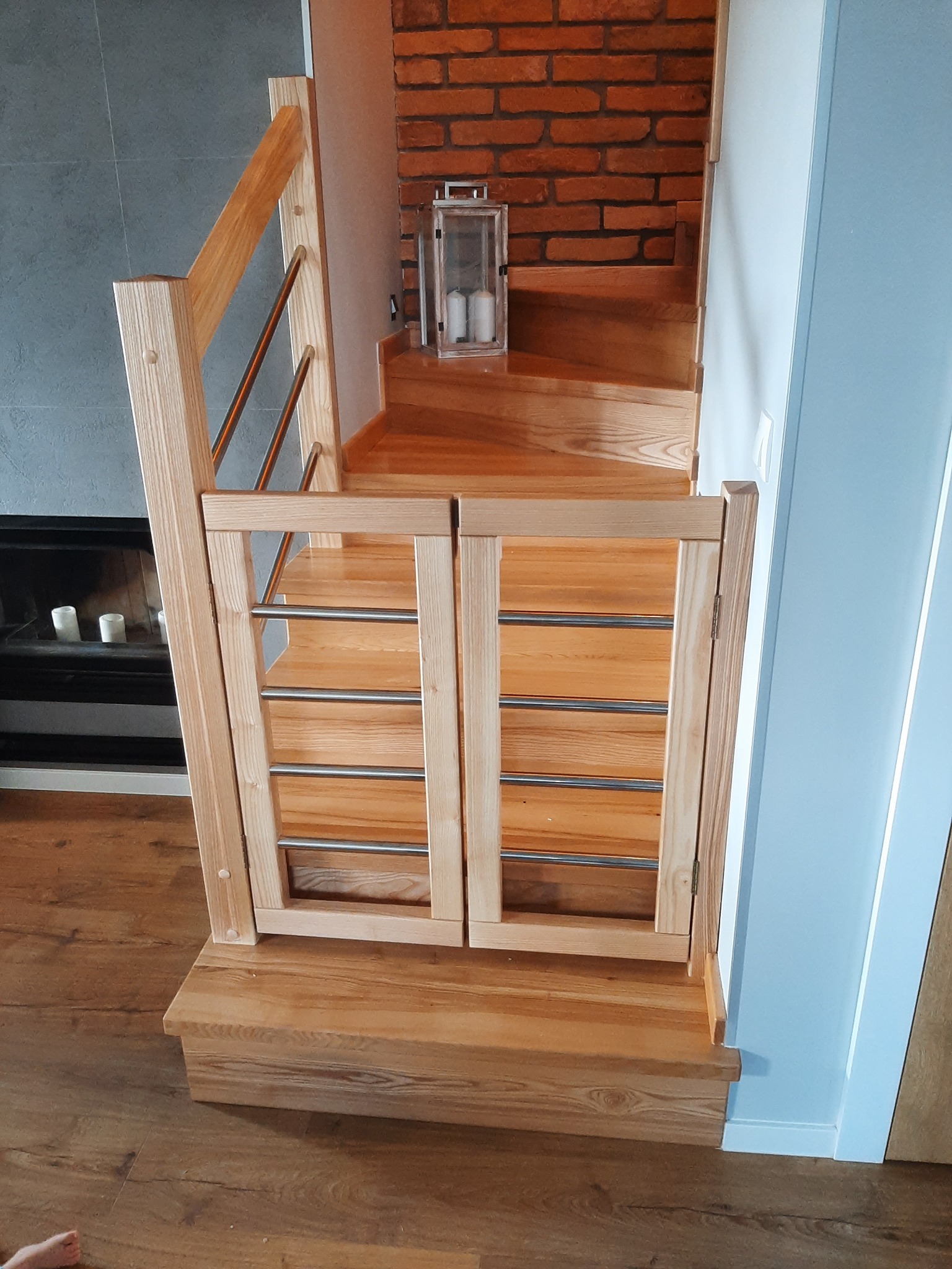 Drewniane schody z zabezpieczeniem w postaci bramki z metalowymi prętami, widok na ceglaną ścianę i kominek z palącymi się świecami.