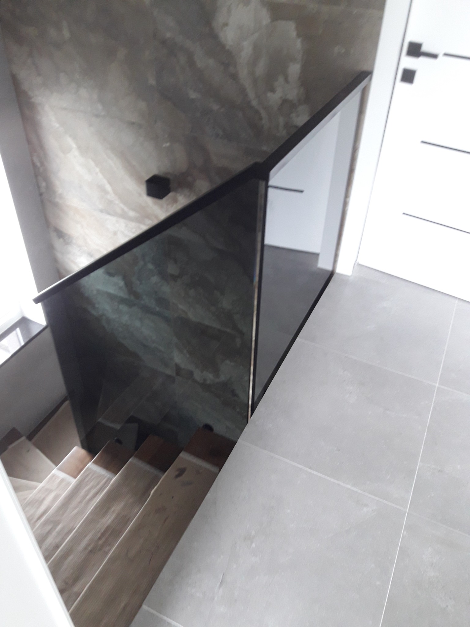 Szklana balustrada z czarnymi profilami zabezpieczająca schody drewniane, widok z góry, ściana z efektem marmuru i białe drzwi w tle.