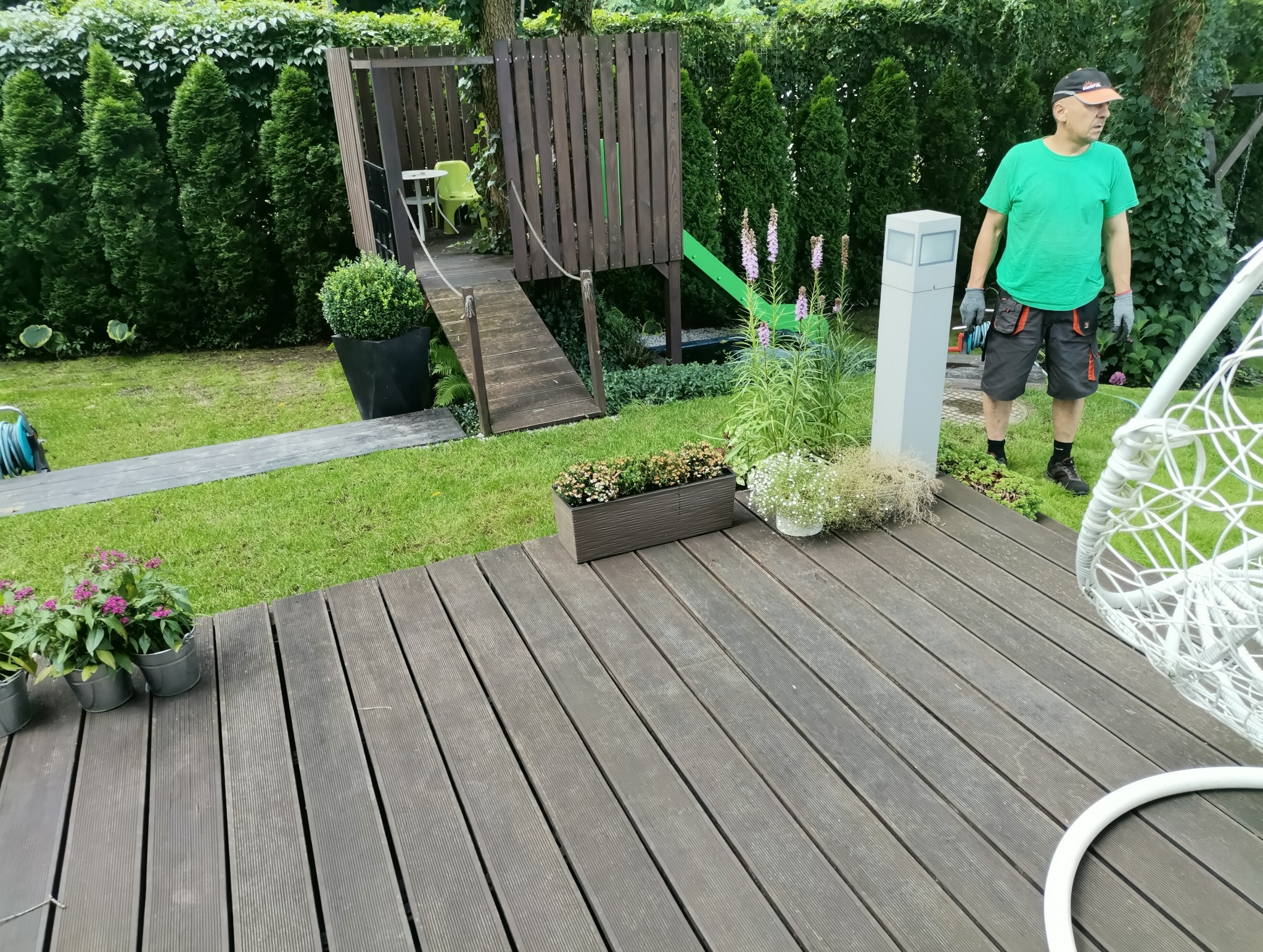 Uporządkowany ogród z drewnianym tarasem, ozdobnymi roślinami w donicach i wiszącym fotelem; w tle pracownik ogrodowy w zielonej koszulce i ogrodowa konstrukcja z mostkiem i zjeżdżalnią.