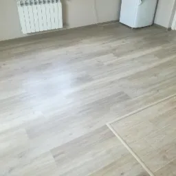 Jasne panele podłogowe ułożone w pomieszczeniu z białym grzejnikiem i lodówką, widoczny fragment progu.