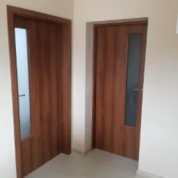 Dwa brązowe drzwi wewnętrzne z szybami, zamontowane w pomalowanym na beżowo korytarzu z marmurową podłogą.