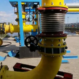 Żółte rurociągi przemysłowe z kompensatorem i zaworami, na tle błękitnego nieba i zabudowań przemysłowych.