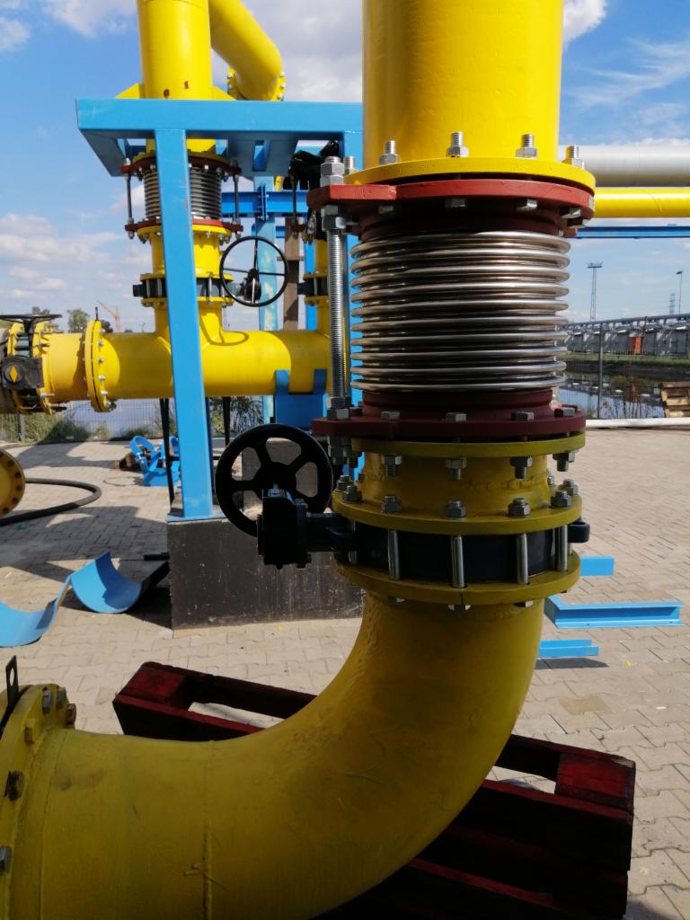 Żółte rurociągi przemysłowe z kompensatorem i zaworami, na tle błękitnego nieba i zabudowań przemysłowych.