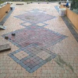 Niedokończona nawierzchnia z kostki brukowej w geometryczny wzór rombów z szarym obramowaniem, widoczne narzędzia i materiały budowlane w tle, pochmurna pogoda.