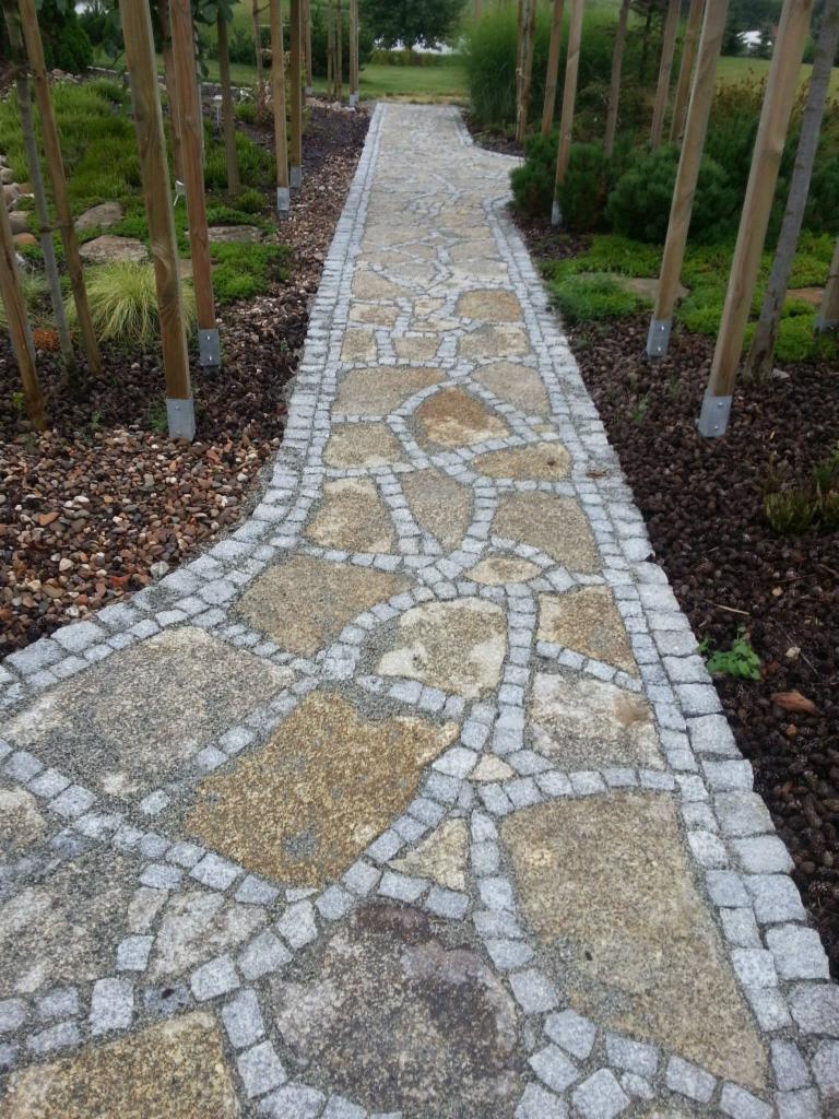 Ścieżka w ogrodzie wyłożona nieregularnymi płytami z jasnego kamienia, obramowana szarą kostką, otoczona młodymi drzewkami i korą.