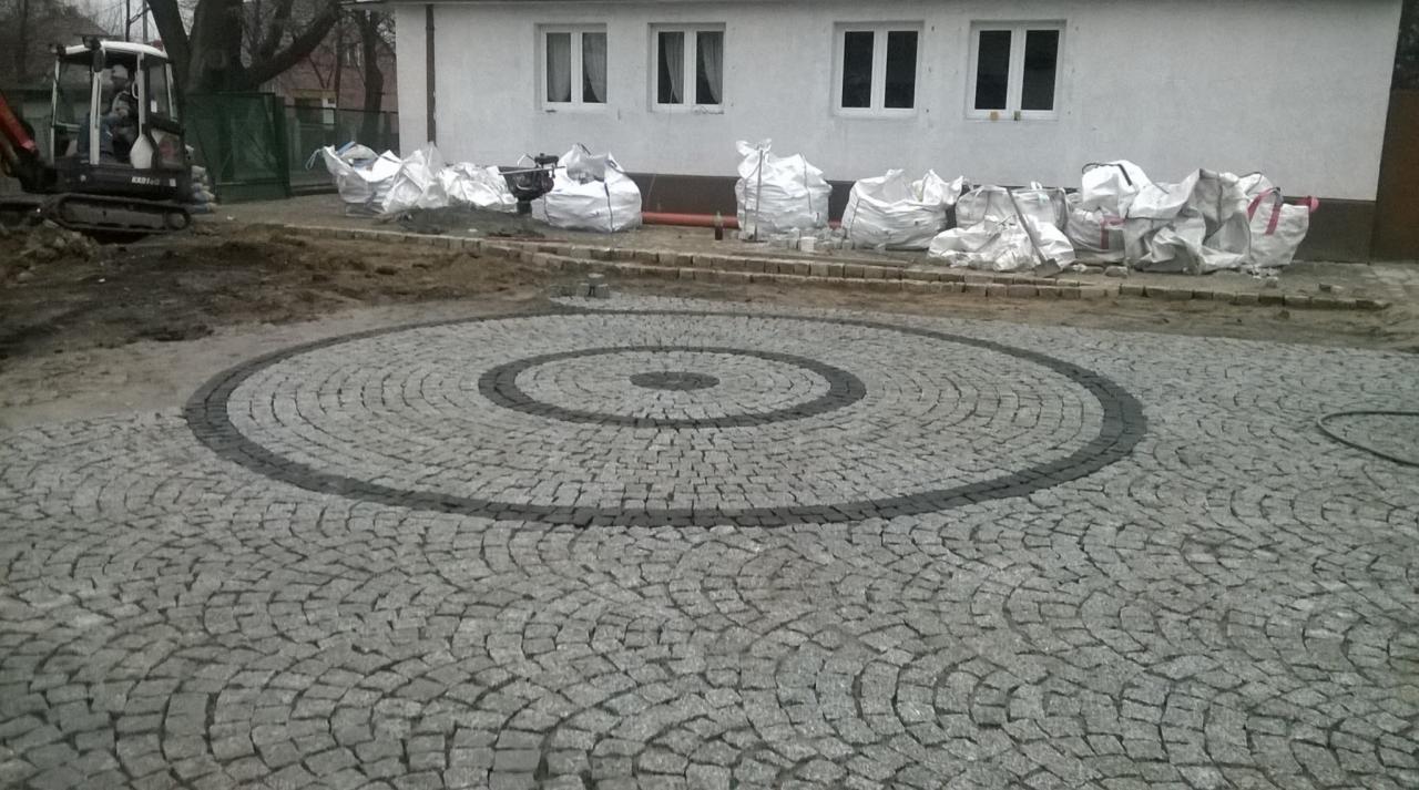 Okazały okrąg z szarej kostki granitowej z ciemniejszymi obwódkami ułożony na podwórku, w tle budynek i worki z materiałem oraz mała koparka.