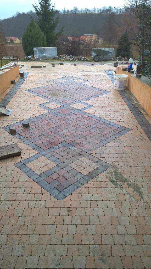 Niedokończona nawierzchnia z kostki brukowej w geometryczny wzór rombów z szarym obramowaniem, widoczne narzędzia i materiały budowlane w tle, pochmurna pogoda.