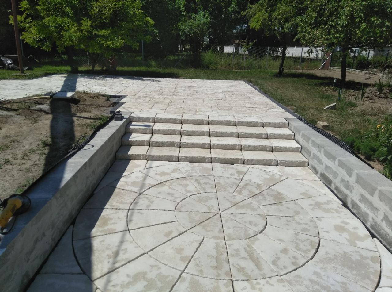 Wykonana z jasnych płyt kamiennych nawierzchnia z okrągłym wzorem i schodami, otoczona niskim murem z betonowych bloczków, w tle zieleń ogrodu.