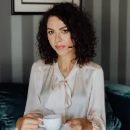 Kobieta z kręconymi włosami siedzi na szmaragdowej sofie, trzymając białą filiżankę z kawą, ubrana w bluzkę z kokardą i bordową spódnicę, w tle obraz w czarnej ramie i szare pionowe pasy.