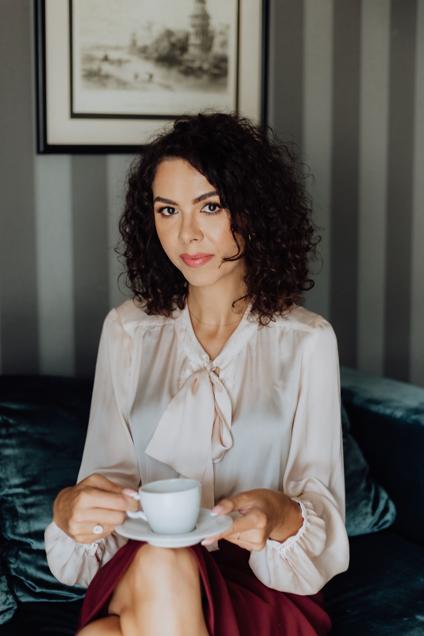 Kobieta z kręconymi włosami siedzi na szmaragdowej sofie, trzymając białą filiżankę z kawą, ubrana w bluzkę z kokardą i bordową spódnicę, w tle obraz w czarnej ramie i szare pionowe pasy.