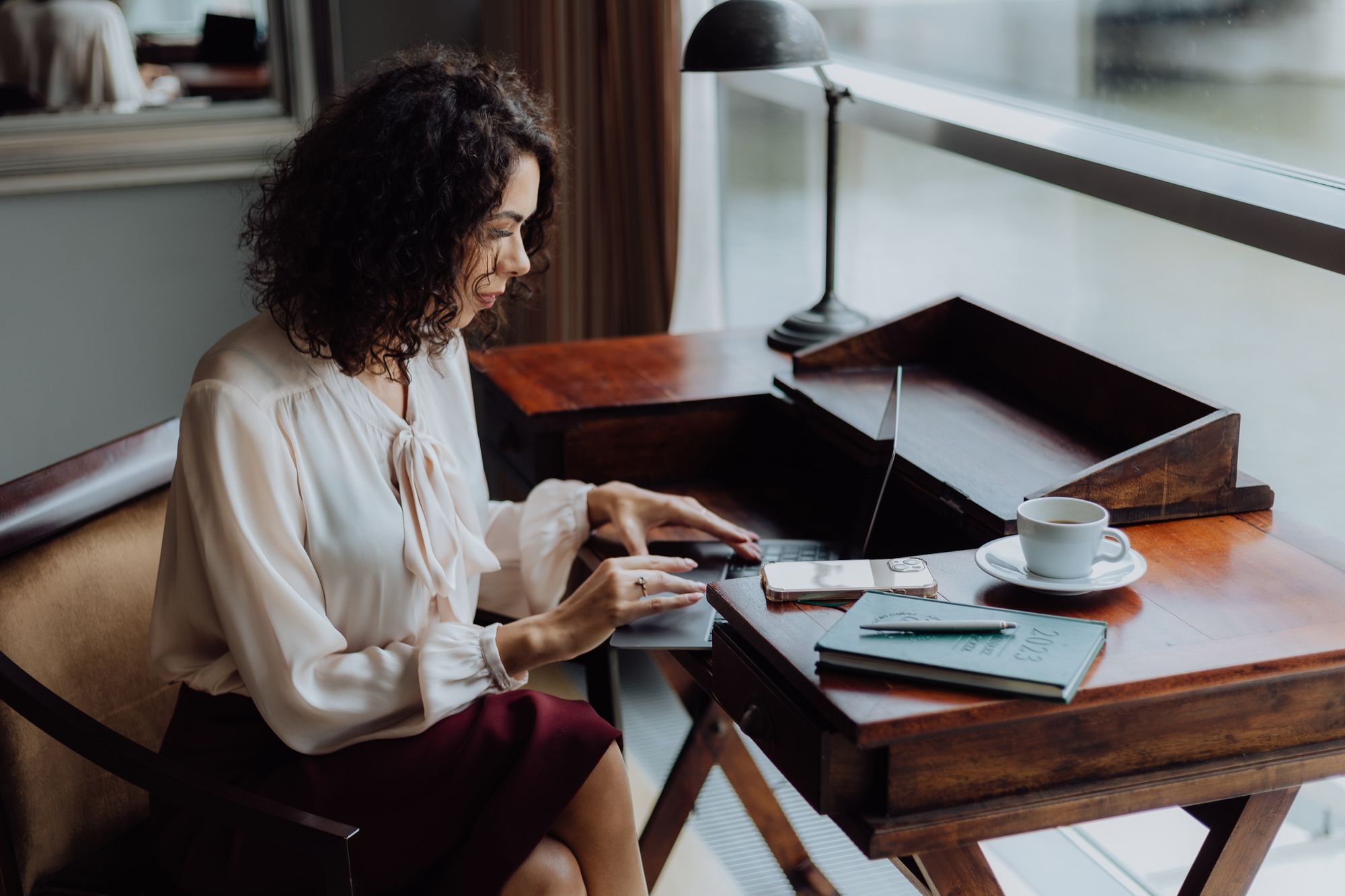 Kobieta z kręconymi włosami pracuje na laptopie przy drewnianym biurku w jasnym pomieszczeniu, obok filiżanka kawy, telefon i kalendarz 2023.