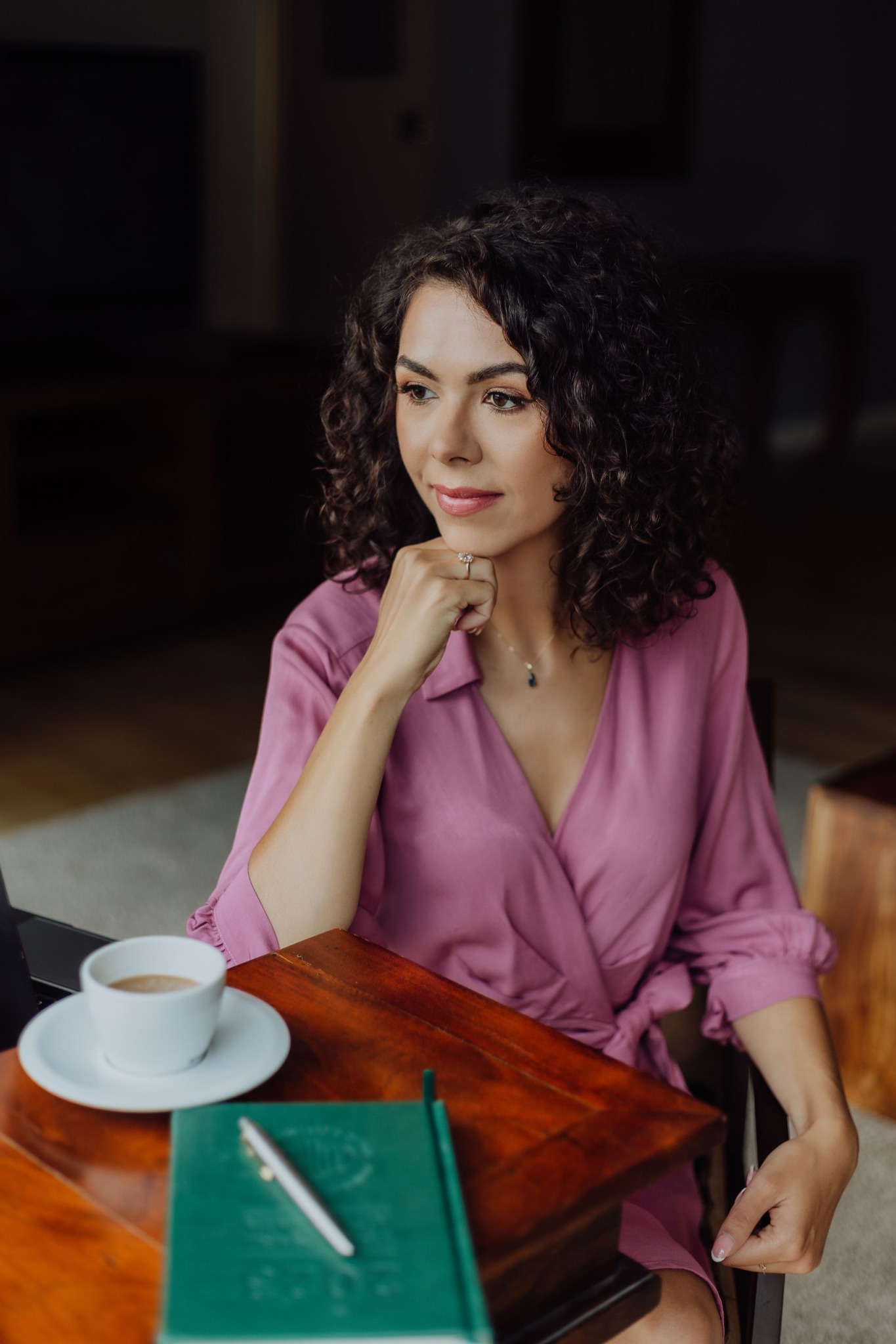 Kobieta z kręconymi włosami, w fioletowej sukience, z pierścionkiem, zamyślona siedzi przy drewnianym stole z zielonym notesem, długopisem i filiżanką kawy.