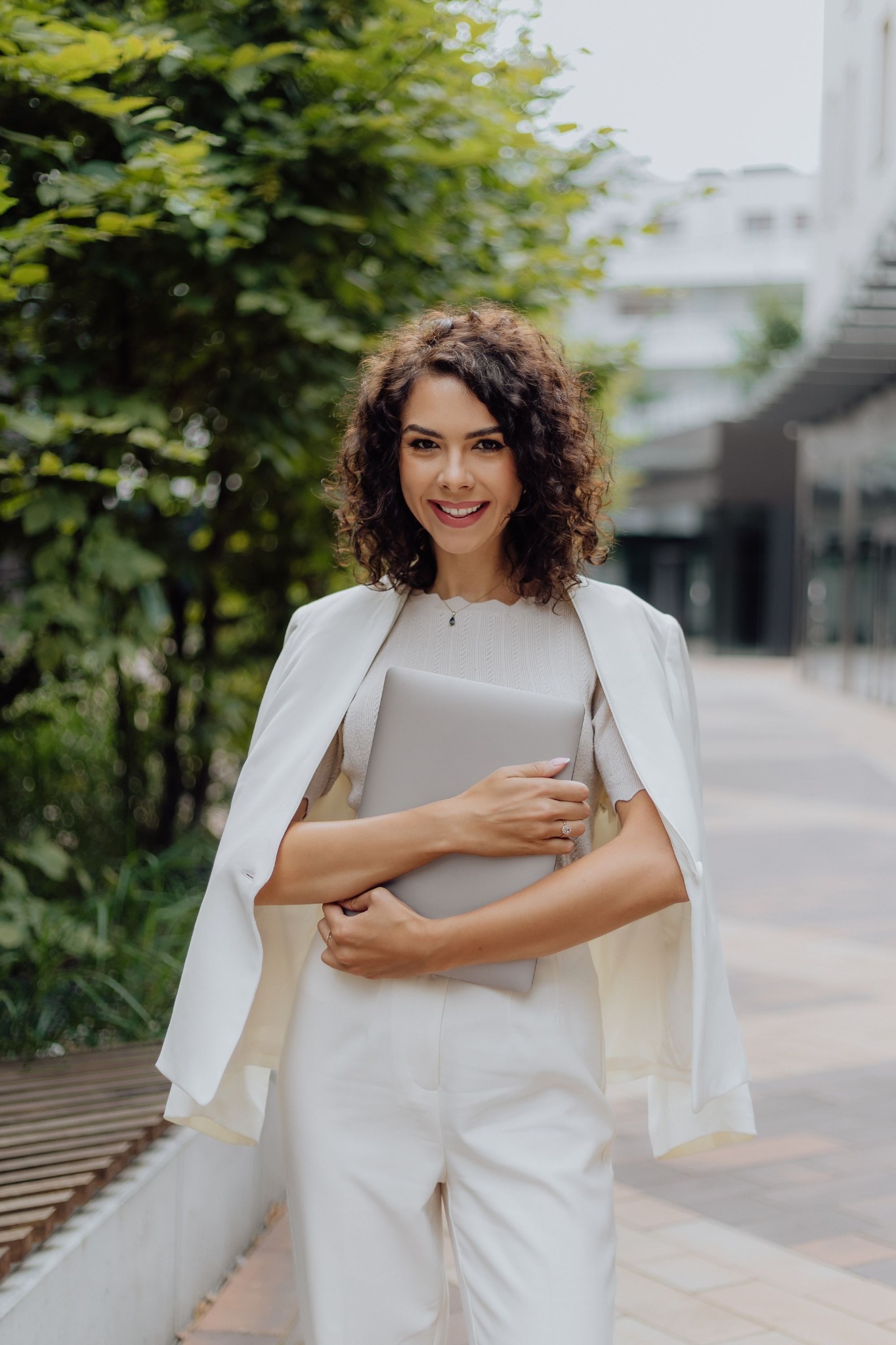 Uśmiechnięta kobieta w białym garniturze, z marynarką narzuconą na ramiona, trzyma zamkniętego laptopa na tle zieleni miejskiej i nowoczesnego budynku.