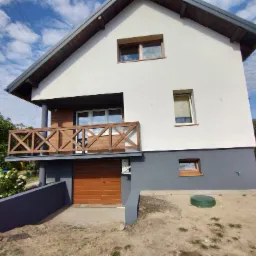 Dwukondygnacyjny dom z balkonem o drewnianej balustradzie, świeżo pomalowaną elewacją w kolorze białym i szarym, widok z dołu na tle błękitnego nieba z chmurami.