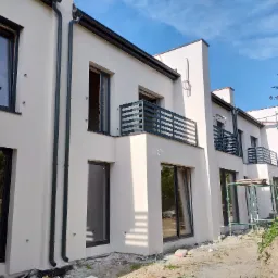 Nowoczesna elewacja budynku wielorodzinnego w jasnym odcieniu, z czarnymi ramami okiennymi i balkonami. Częściowo widoczne rusztowania.