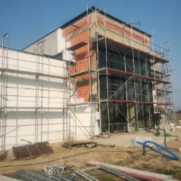 Budynek w trakcie ocieplania, otoczony rusztowaniami. Widoczne fragmenty elewacji z czerwonej cegły oraz biały styropian. Na ziemi leżą elementy rusztowania.