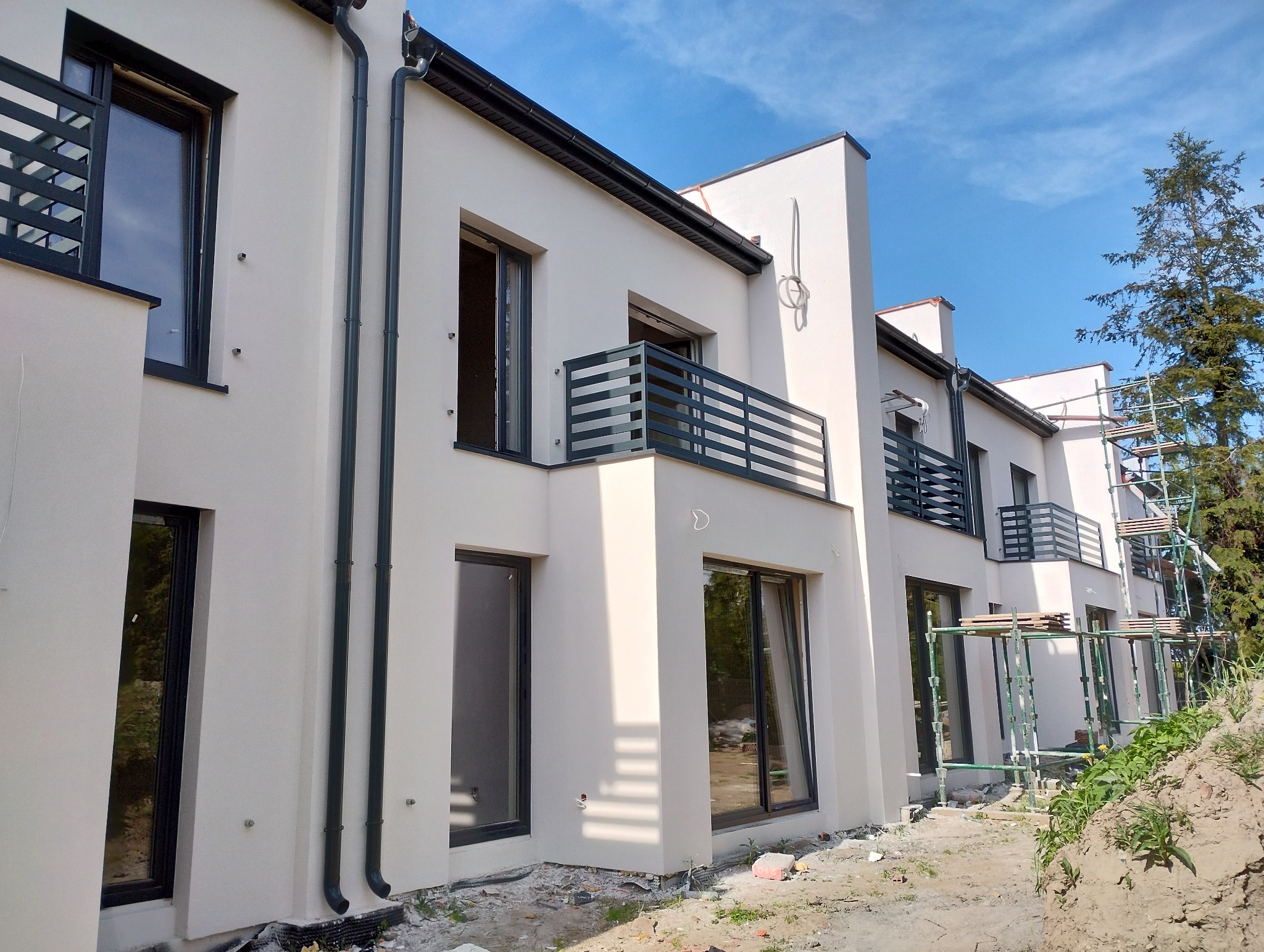 Nowoczesna elewacja budynku wielorodzinnego w jasnym odcieniu, z czarnymi ramami okiennymi i balkonami. Częściowo widoczne rusztowania.