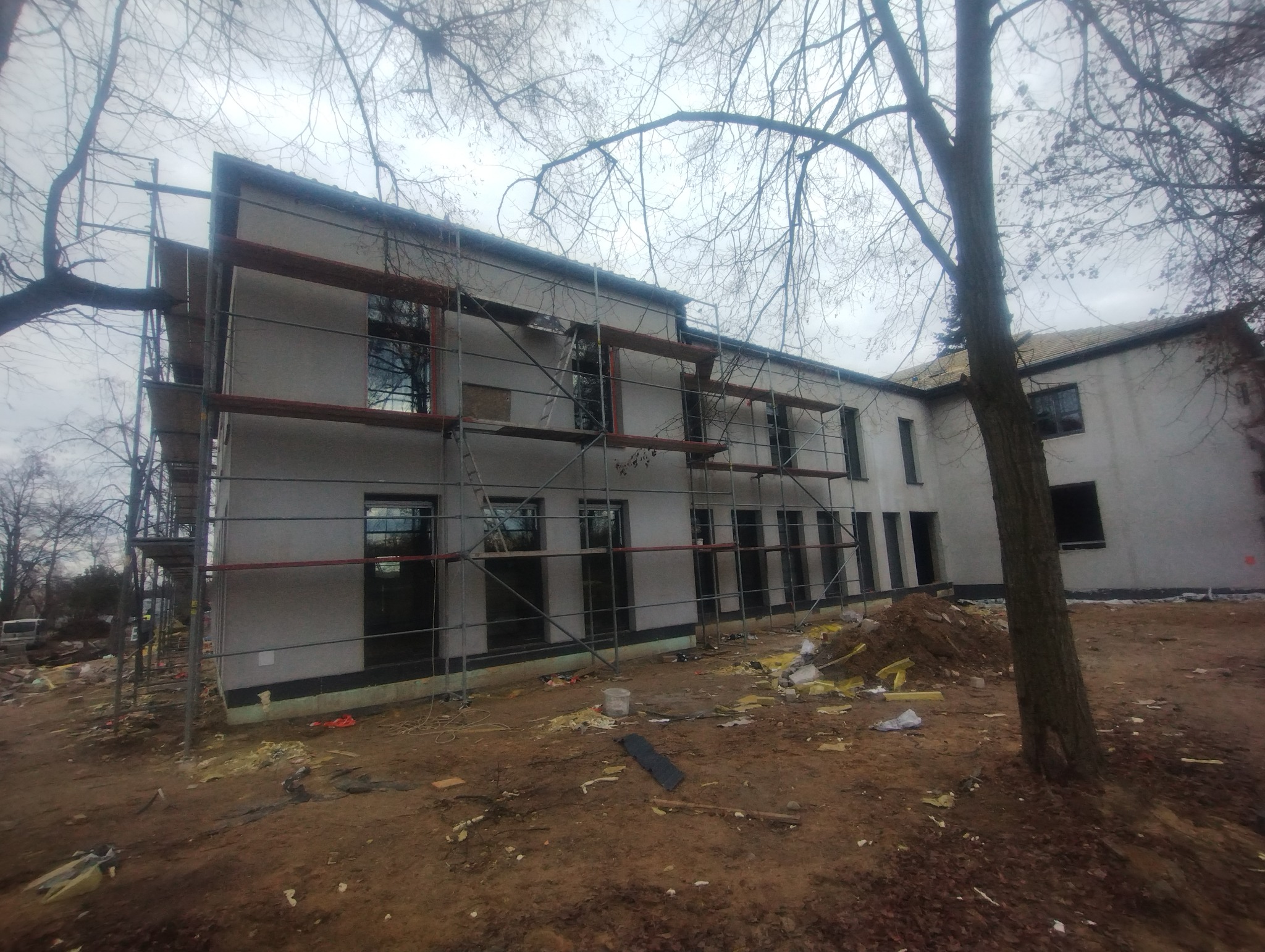 Budynek dwukondygnacyjny w trakcie prac elewacyjnych, otoczony rusztowaniami i z widocznym ociepleniem u podstawy. Na pierwszym planie nieporządek budowlany.