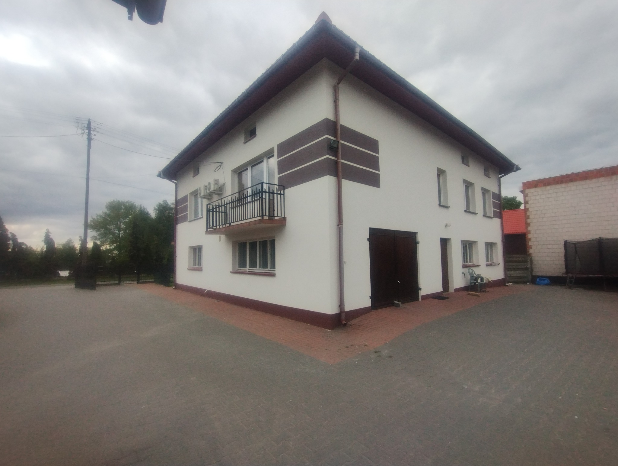 Dwupiętrowy dom z balkonem, białą elewacją i brązowymi akcentami, widoczny z zewnątrz na tle pochmurnego nieba i zieleni. Widoczny fragment kostki brukowej.
