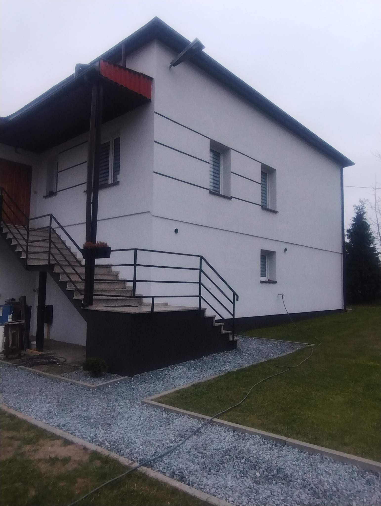 Dwupiętrowy dom z białą elewacją, czarnymi poziomymi pasami i schodami zewnętrznymi. Otoczenie: trawnik i ścieżka z kamieni. Pochmurne niebo w tle.