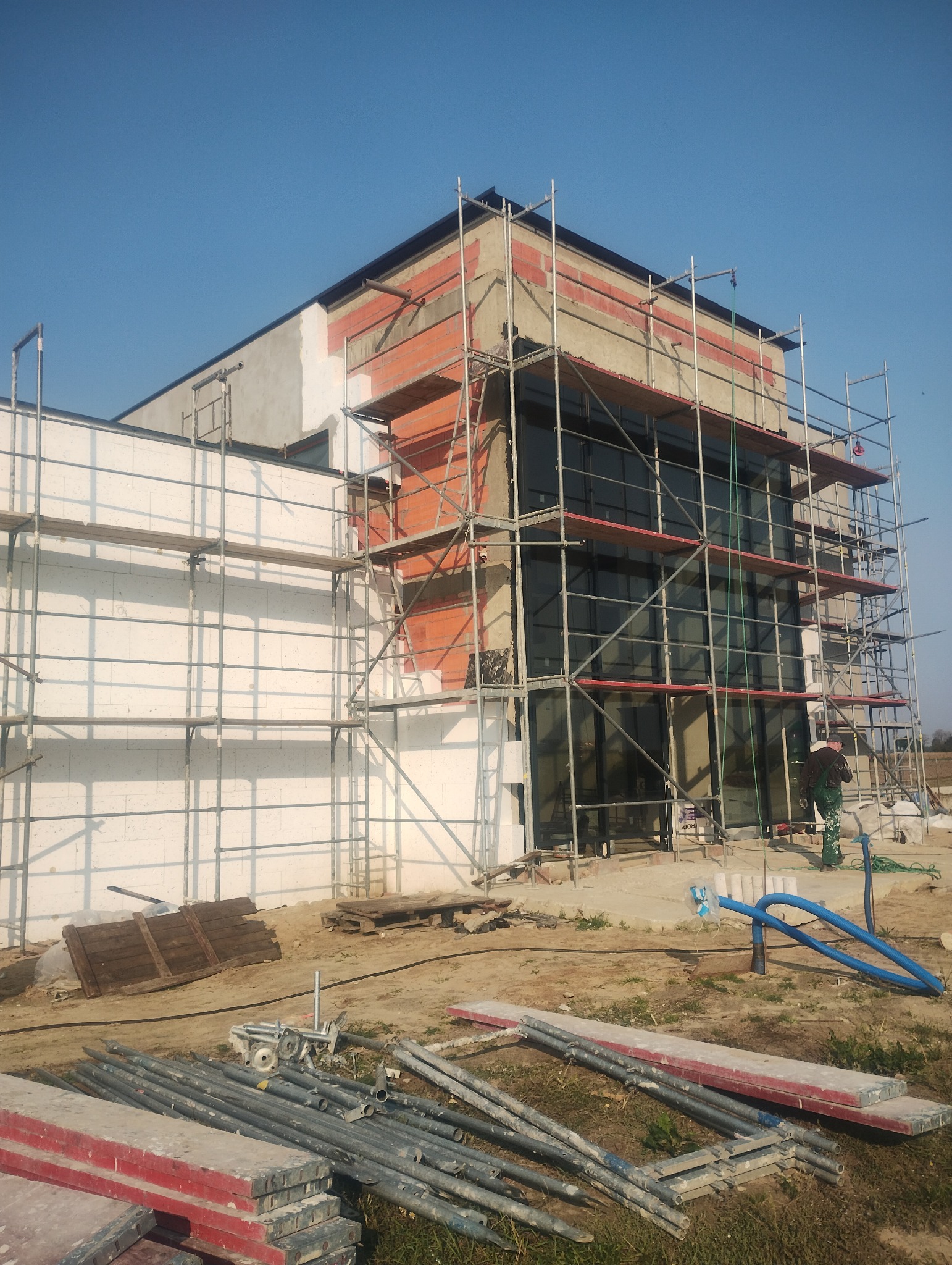 Budynek w trakcie ocieplania, otoczony rusztowaniami. Widoczne fragmenty elewacji z czerwonej cegły oraz biały styropian. Na ziemi leżą elementy rusztowania.