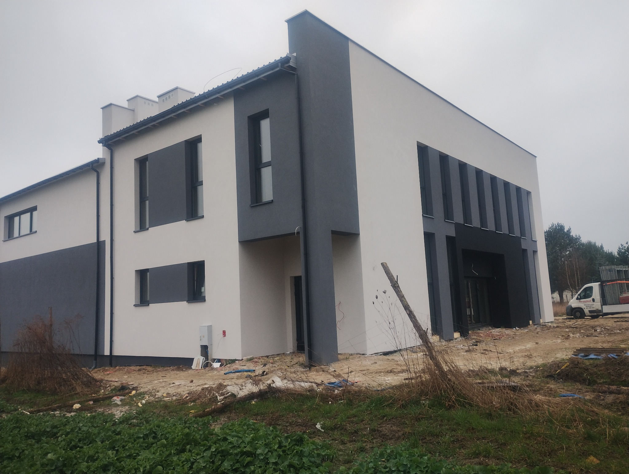 Nowoczesny dom po tynkowaniu i malowaniu elewacji w odcieniach bieli i szarości, widok z zewnątrz, teren wokół domu w trakcie prac wykończeniowych.