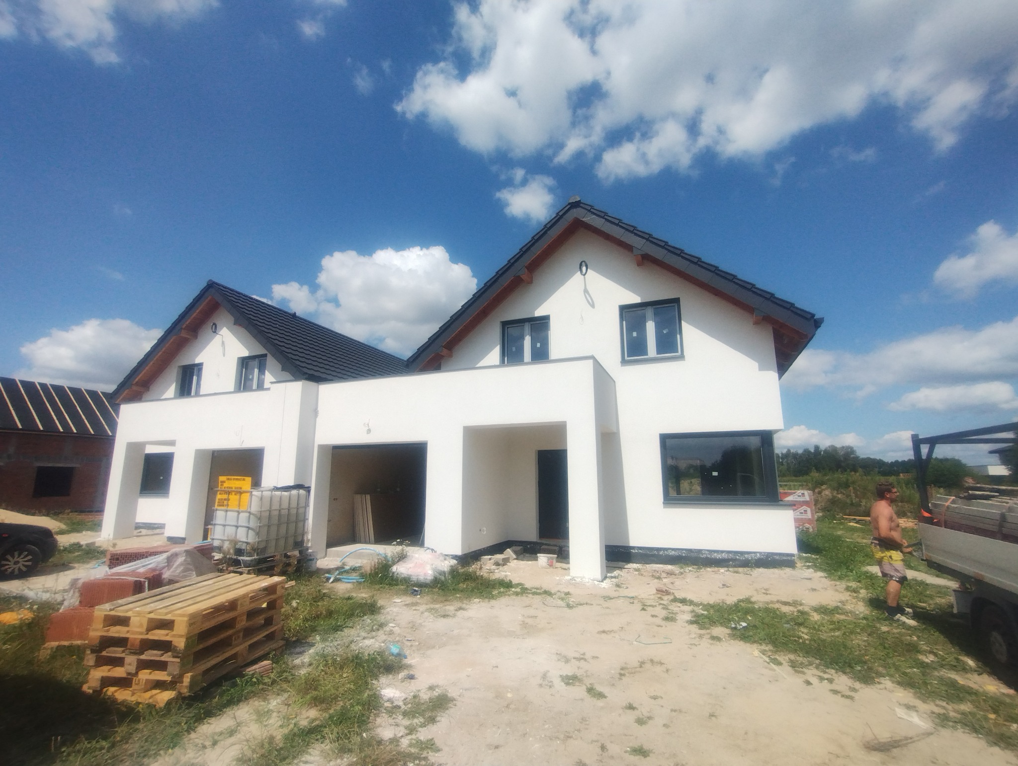 Biały, nowoczesny bliźniak z ciemną dachówką w słoneczny dzień. Widoczne palety i materiały budowlane, sugerujące etap wykończeniowy elewacji.