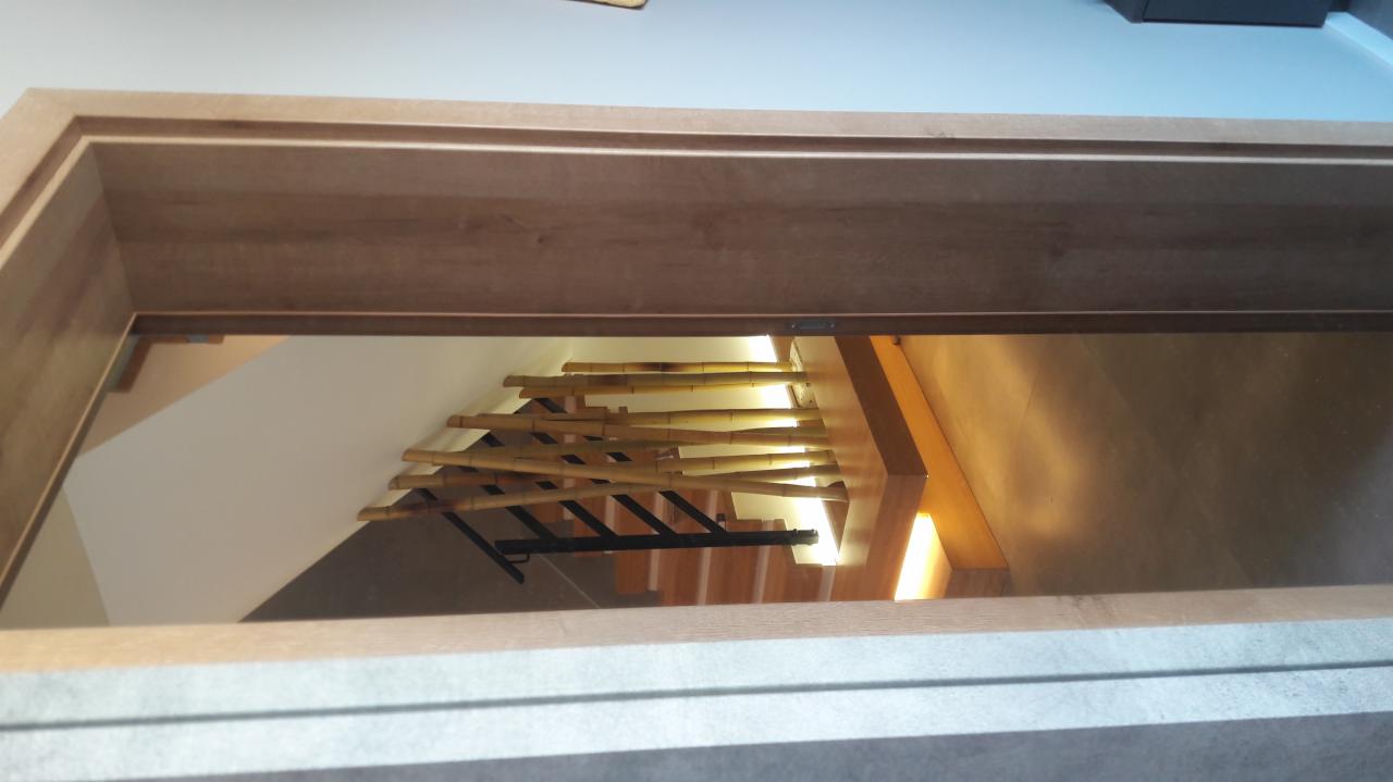 Widok z góry na schody z drewnianymi stopniami i balustradą z bambusowych tyczek, oświetlone od dołu, widziane przez drewnianą ramę drzwi.