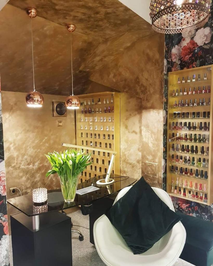 Eleganckie wnętrze salonu kosmetycznego w Zembrzycach z dekoracyjną ścianą, szklanym biurkiem, białym fotelem, bukietem tulipanów i podświetlanymi półkami z lakierami do paznokci.